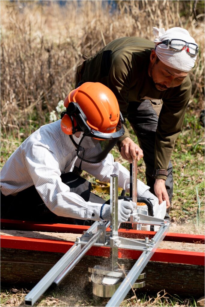 屋外で木を板にする機械を使う木こり隊と指導する木こりの前田さん